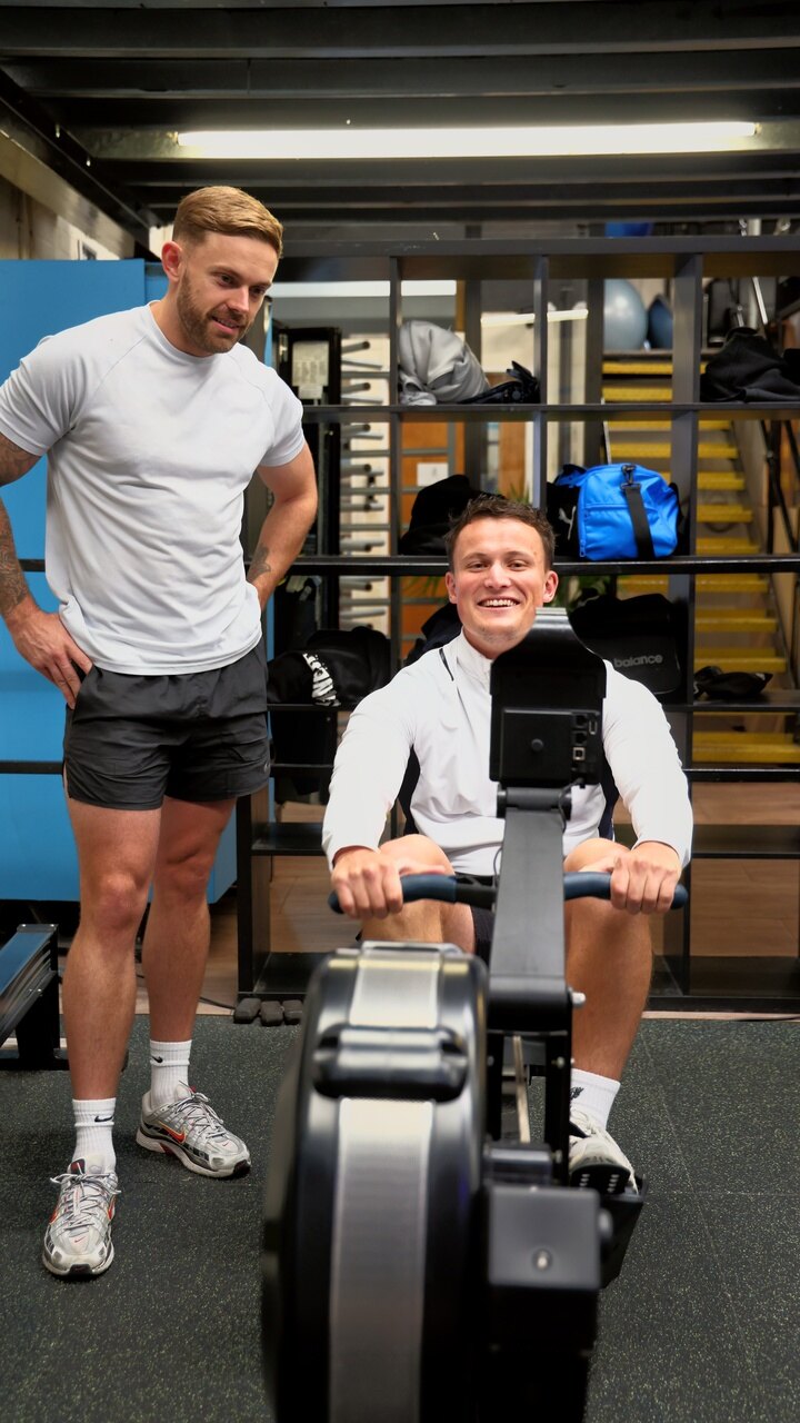 A coach stands over a smiling member on the rowing machine