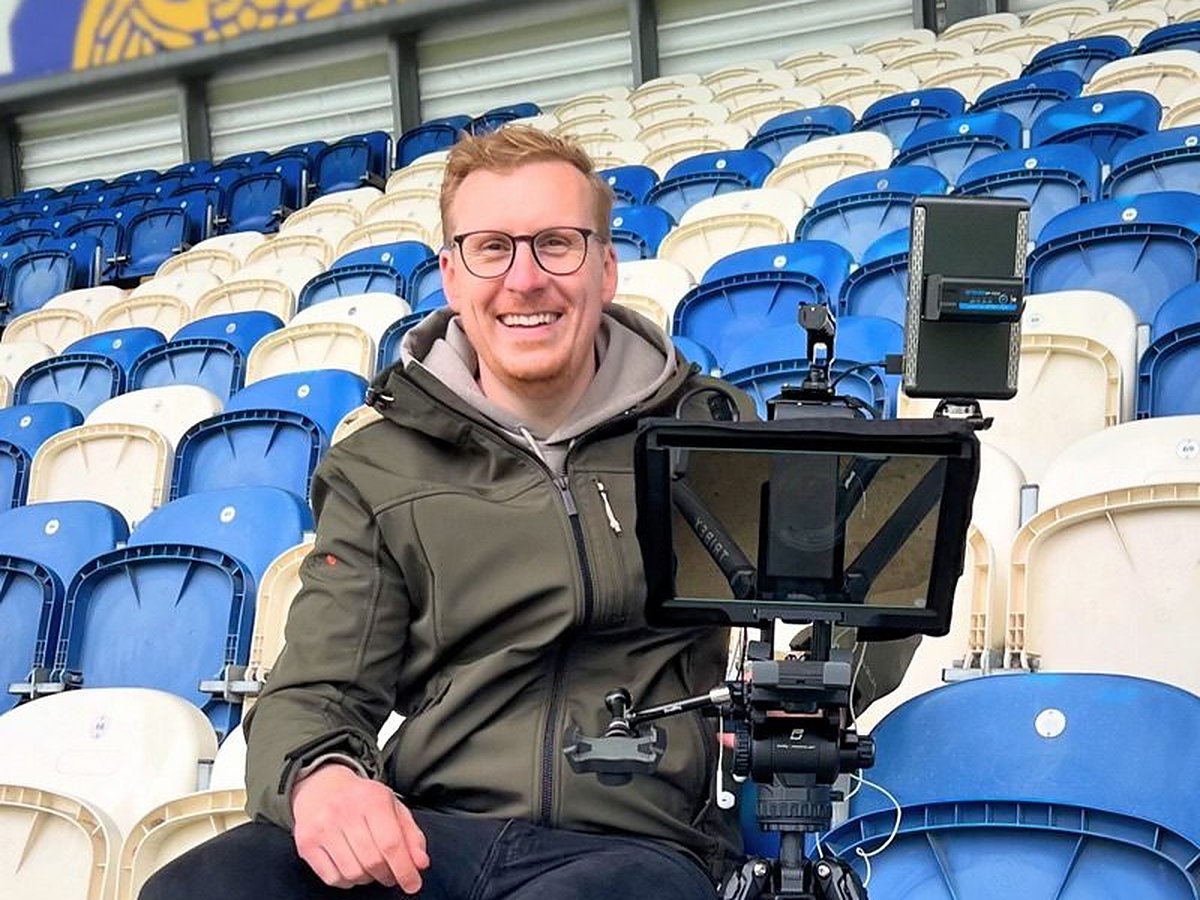 Sam smiling on a stadium seat beside a camera and teleprompter rig on a tripod on a shoot day