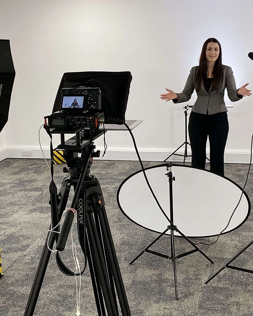 A presenter in a studio set with a camera and teleprompter on a tripod, a softbox and a mic stand
