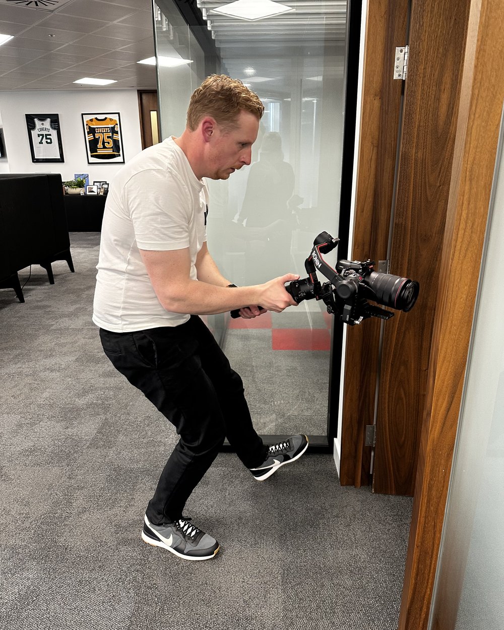 Sam crouching with a gimbal camera through an office doorway to shoot a moving shot