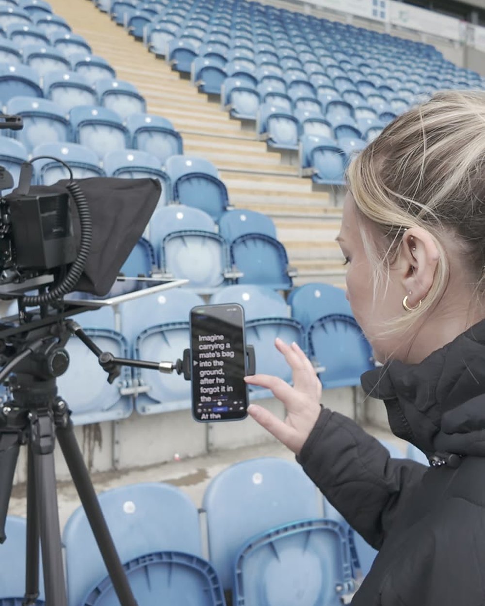 A crew member holding a phone script beside a teleprompter rig on a tripod in a football stadium