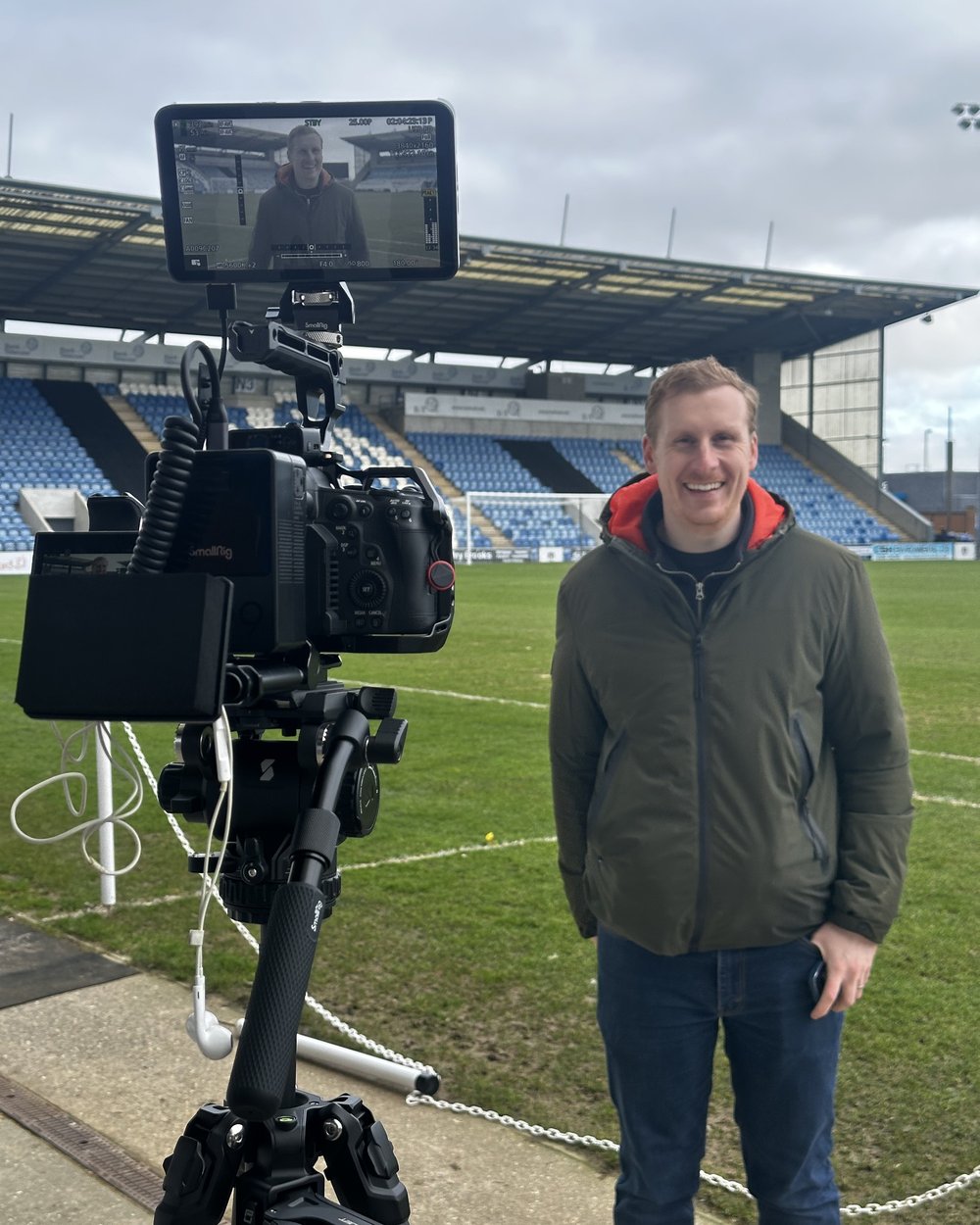 Sam smiling beside a camera and monitor on a tripod at the edge of a football pitch