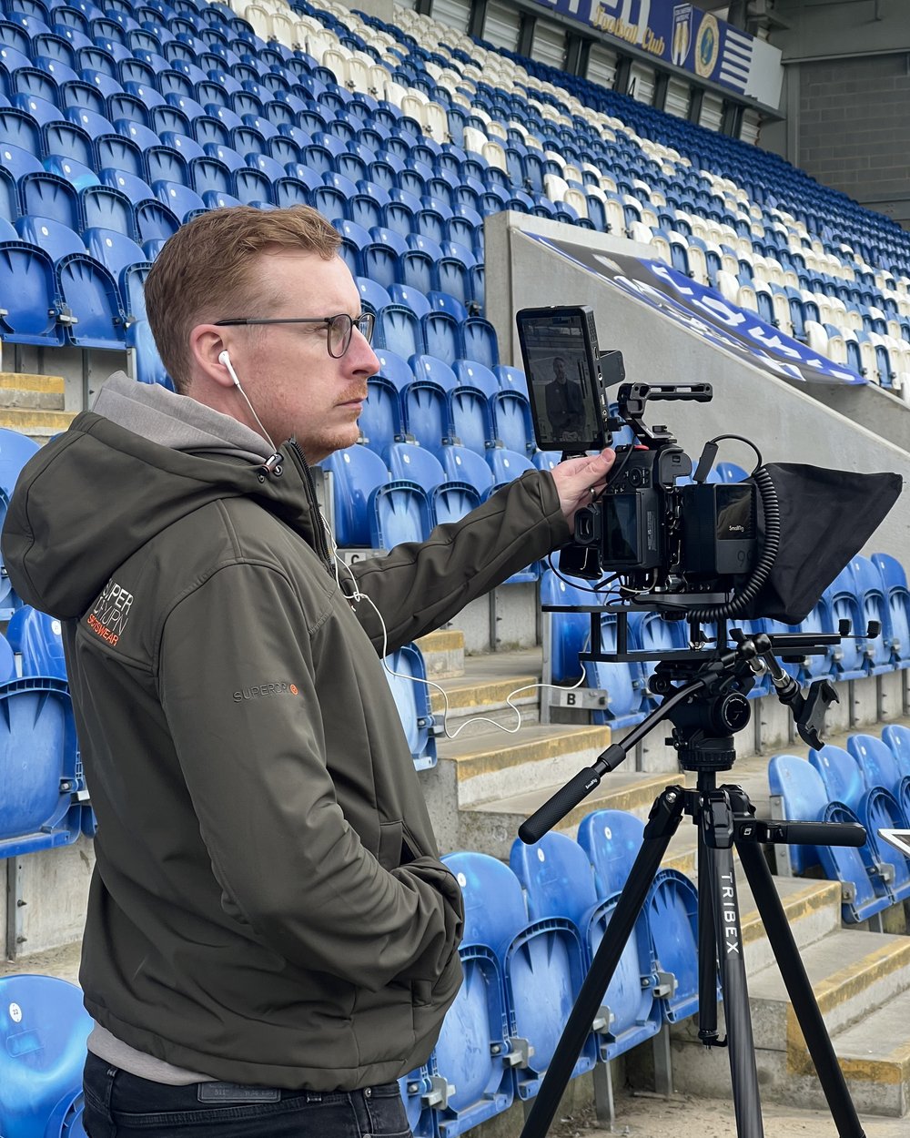 Sam adjusting the teleprompter hood on a camera rig in a football stadium stand