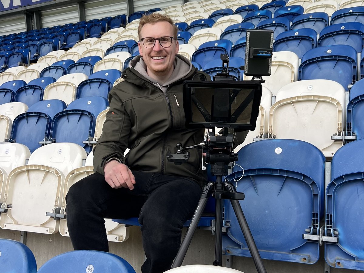 Sam smiling behind a camera and teleprompter on a tripod in a stadium stand