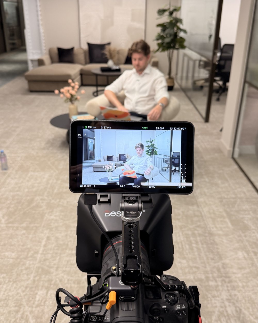 A camera monitor framing a presenter in a furniture showroom lounge, with our rig in the foreground