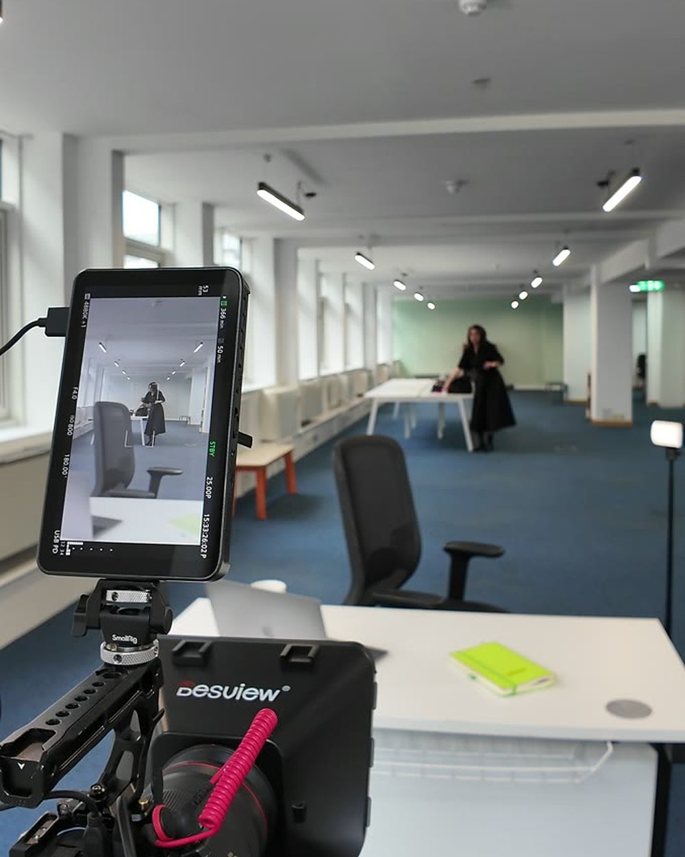 Camera monitor showing a wide shot of an open plan office with a laptop and notebook on the desk