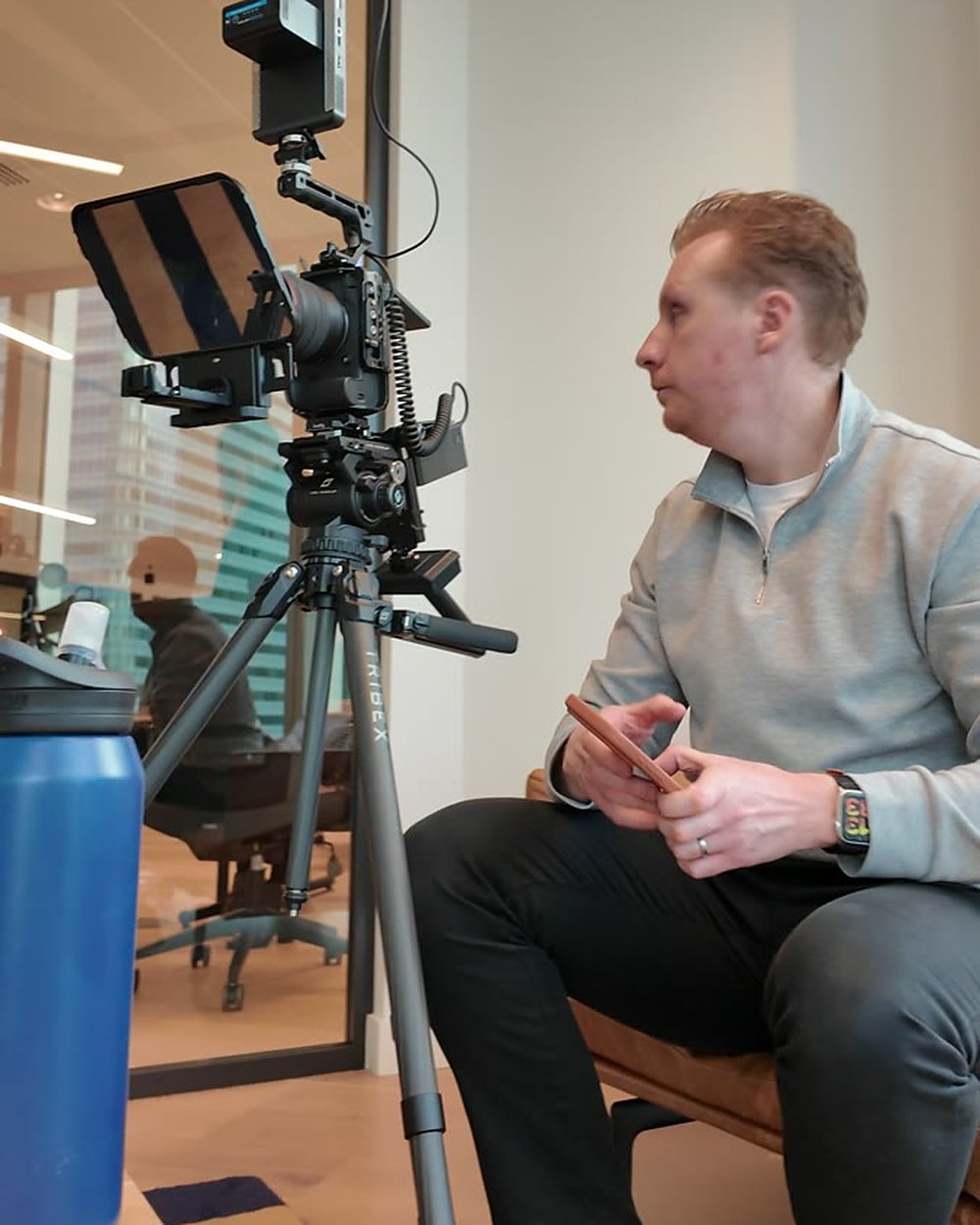 Sam sitting beside a camera and teleprompter between takes in a glass office