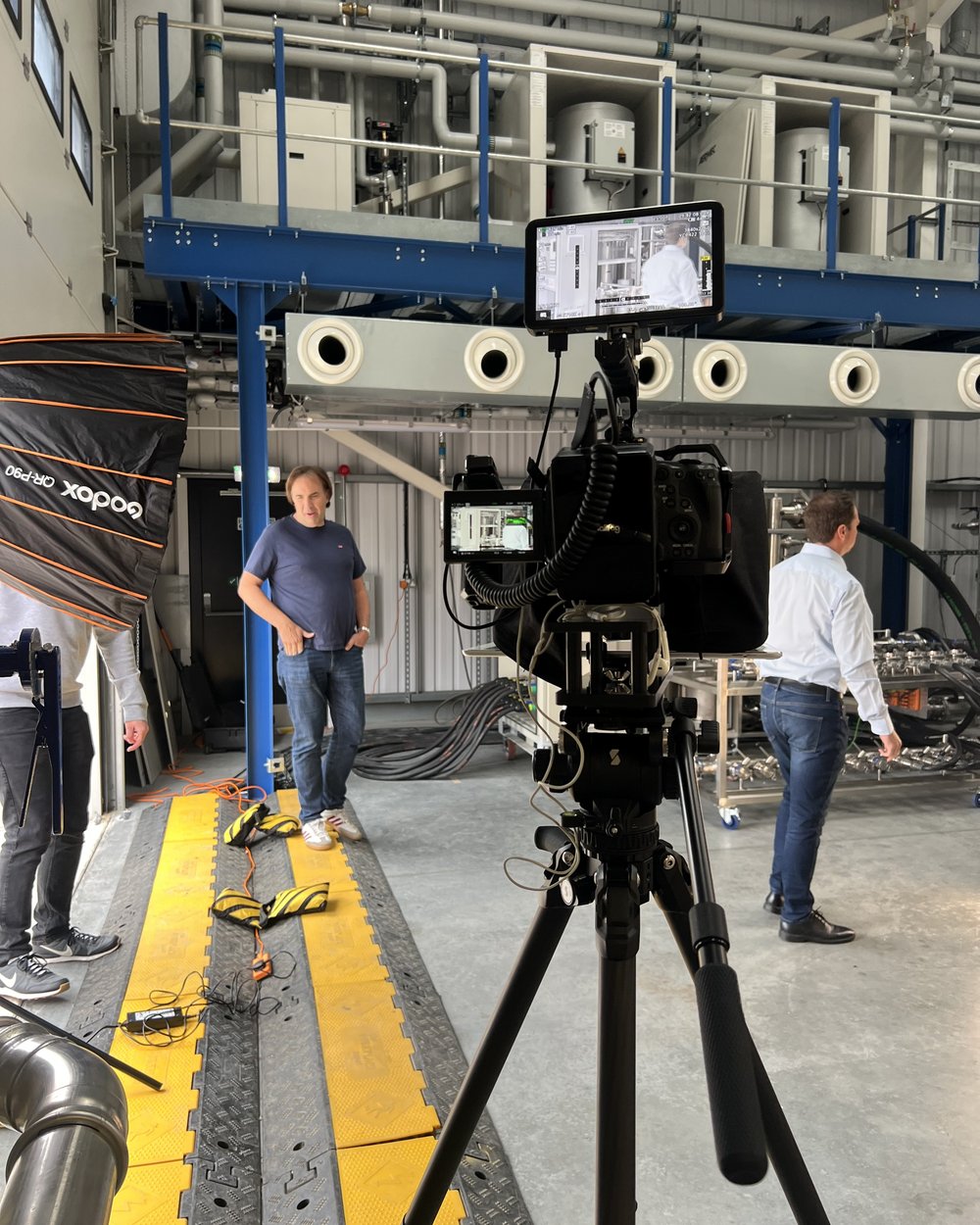 A camera rig with a monitor in the foreground and a softbox lighting a factory floor, the team working behind