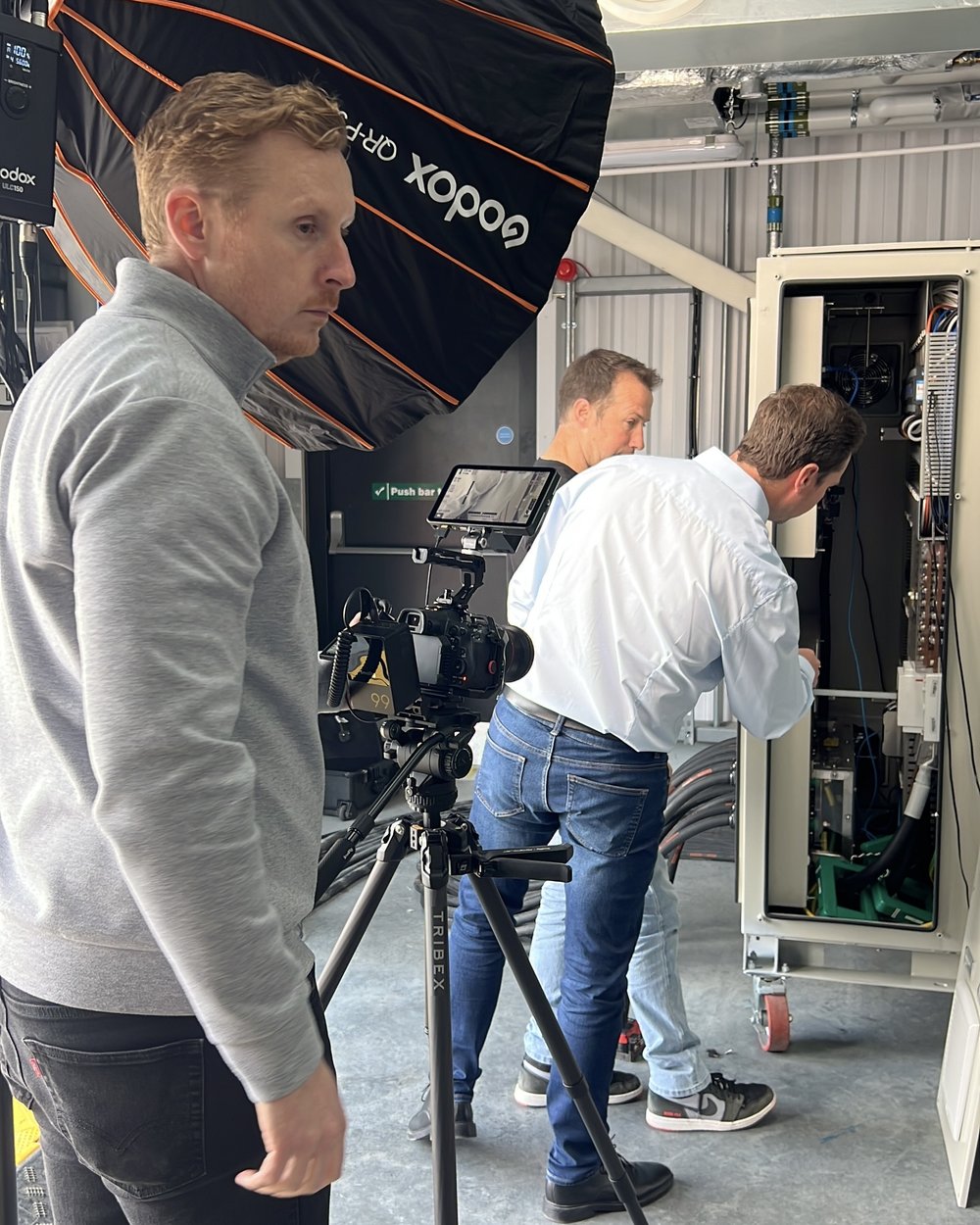 Sam by a softbox light beside a camera on a tripod while a PowerLoad engineer works at an equipment cabinet