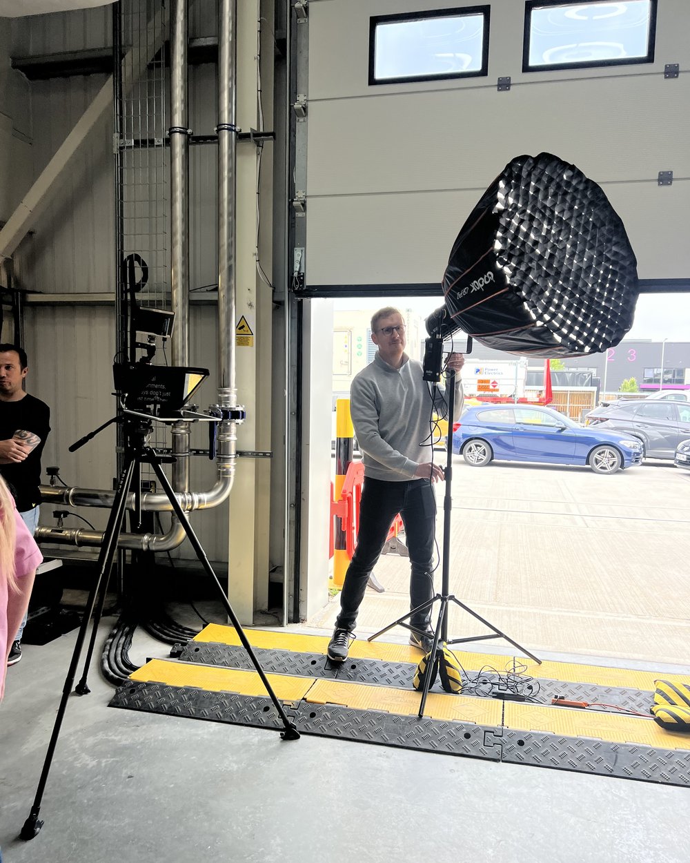 Camera and teleprompter rig with a softbox lighting a factory bay while Sam sets the light