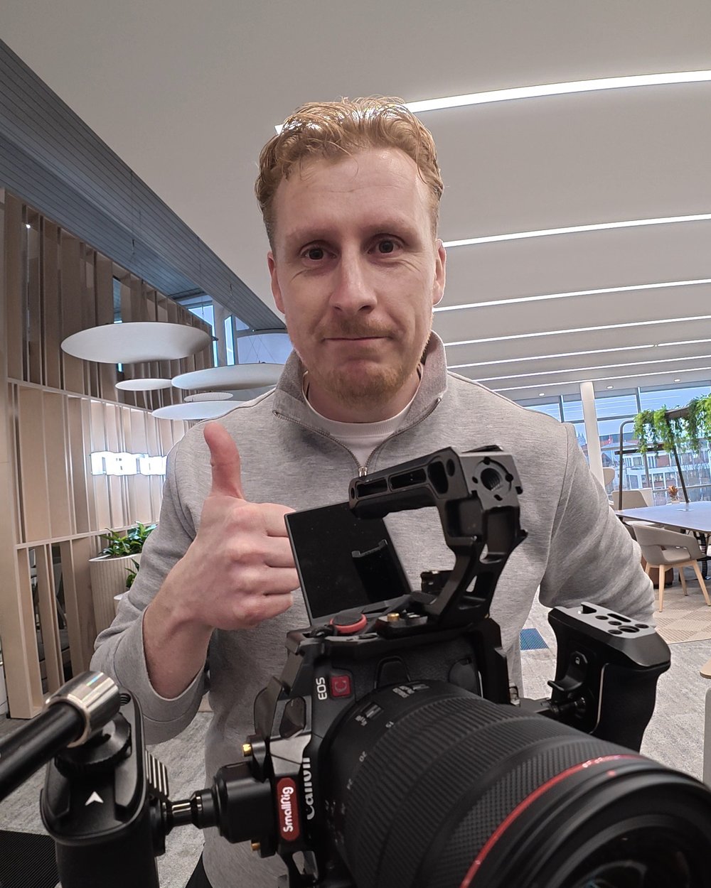 Sam giving a thumbs up behind a cinema camera with a large lens in an open plan office