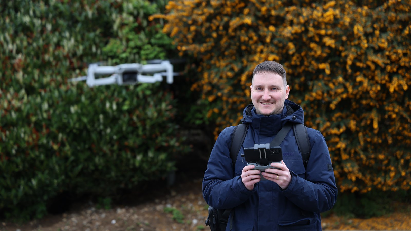 A drone operator holding the controller and smiling while the drone hovers behind him