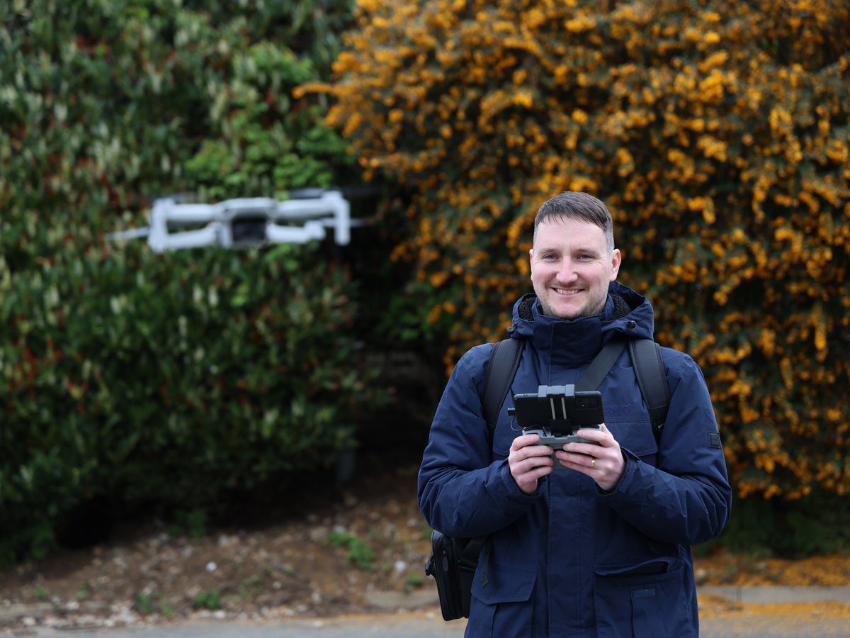 Sam smiling with a drone controller in his hands while the drone hovers behind him outdoors