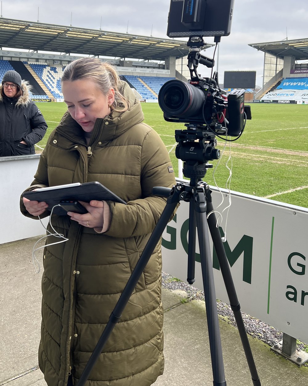 A crew member in a parka reads a script on a tablet beside a cinema camera on a tripod on a football pitch