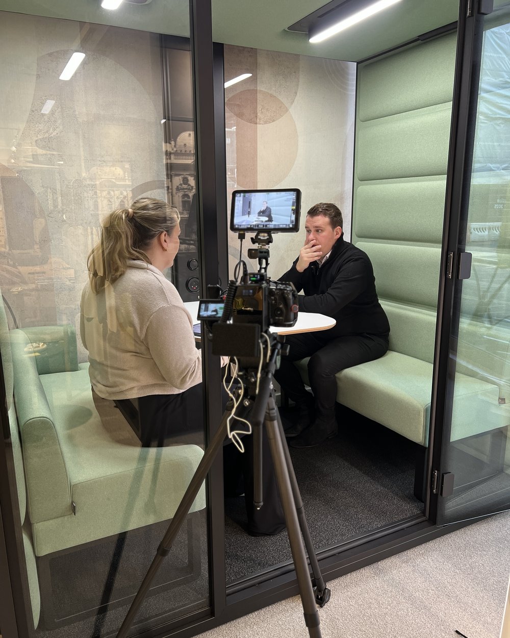 A producer interviewing a team member in a glass meeting pod with the camera and monitor on a tripod