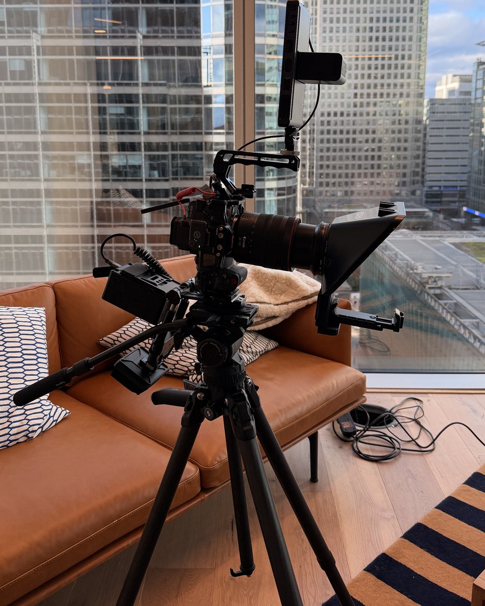 A camera with a teleprompter fitted on a tripod in front of a sofa and city skyline windows