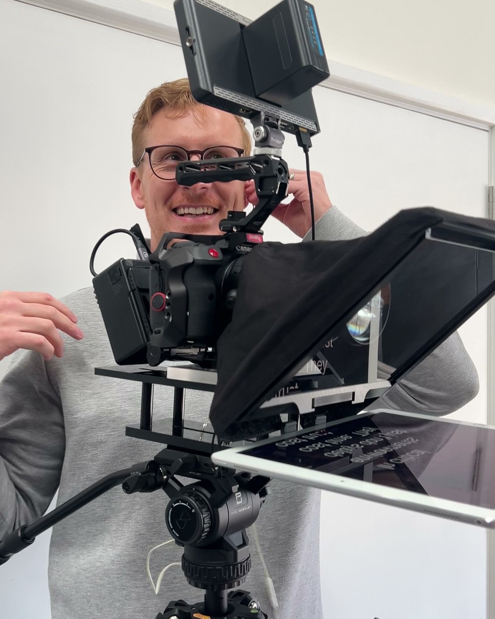 Sam smiling behind a camera fitted with a teleprompter and monitor on a tripod, ready for a take