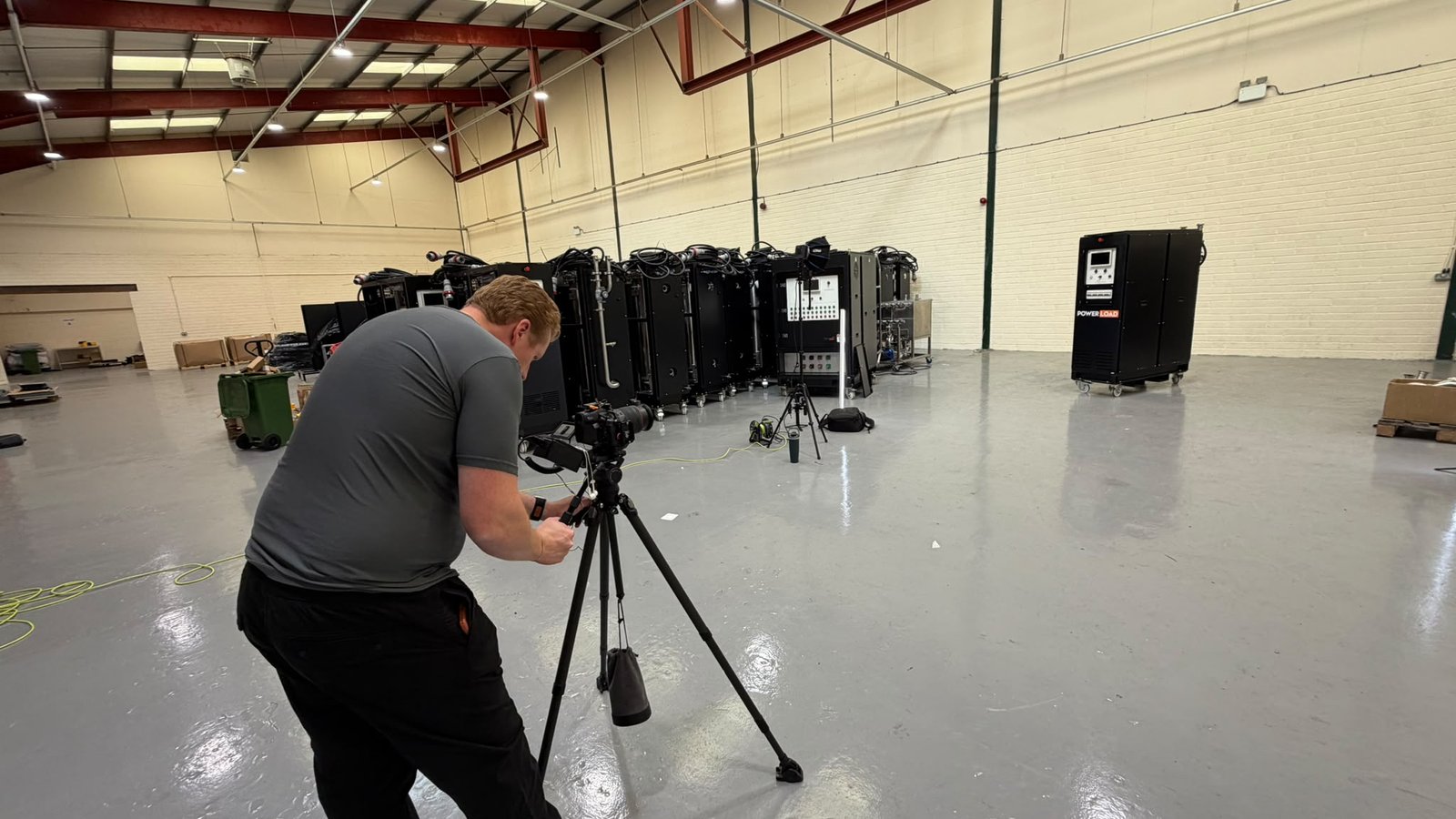 Sam bent over a tripod camera on a factory floor with PowerLoad units lined up behind