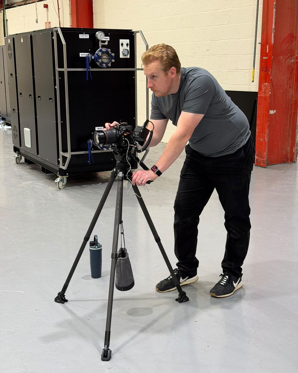 Sam leans over a tripod camera aimed at a rack of PowerLoad cabinets on a clean factory floor