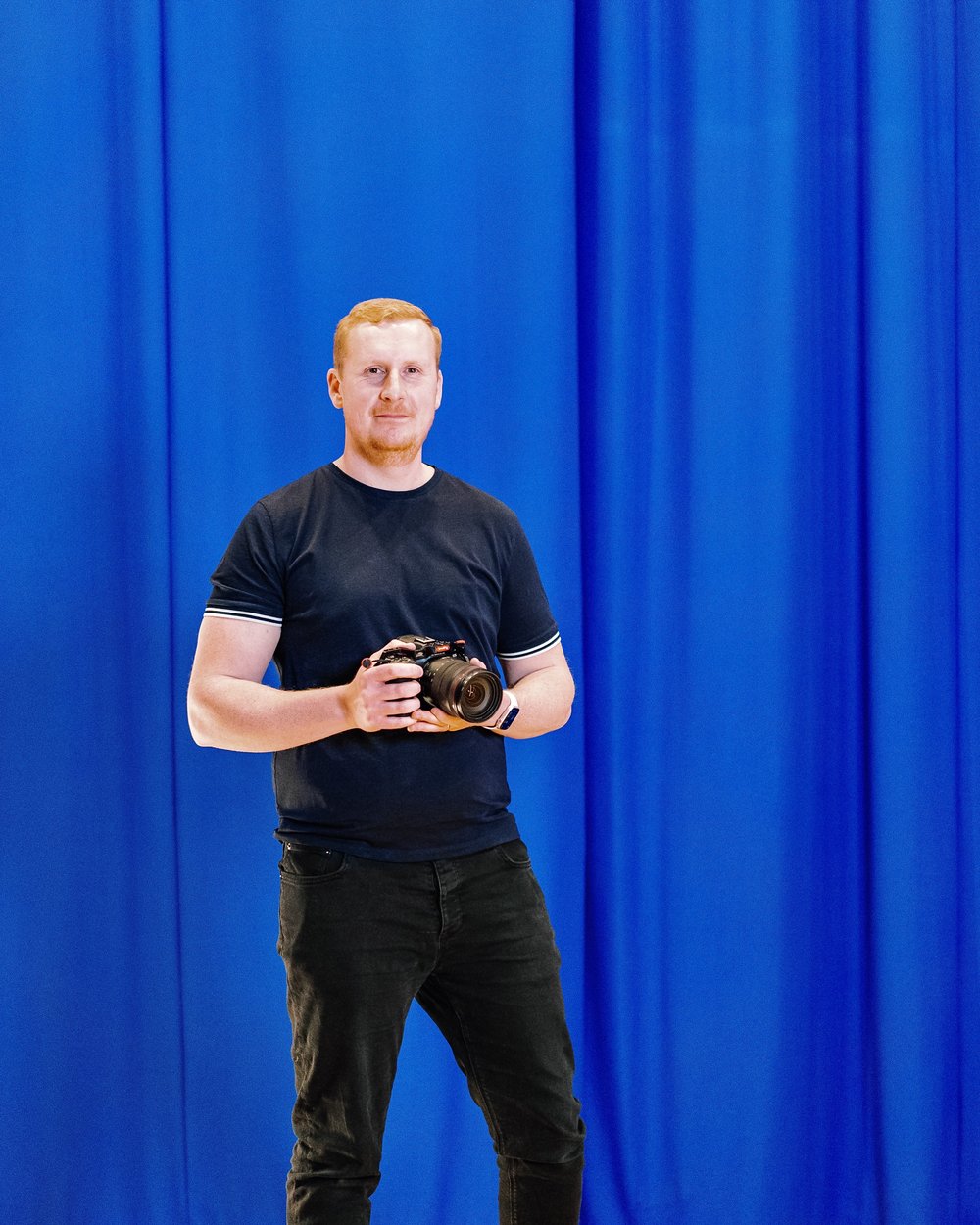 Sam standing in front of a bright blue studio backdrop holding a stills camera, with a light stand behind him