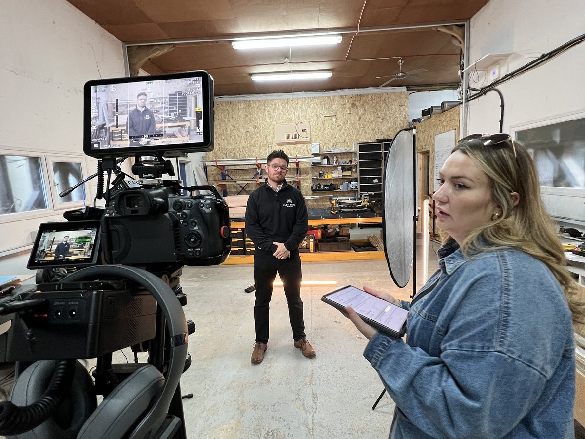 A producer briefs a presenter in a workshop beside a camera and monitor