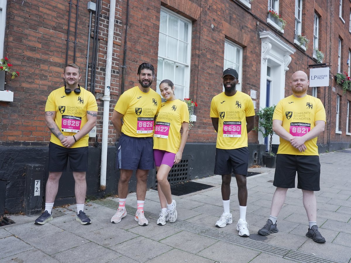 Five team members in matching yellow running tops with race numbers, standing together outside a brick building before Run Norwich