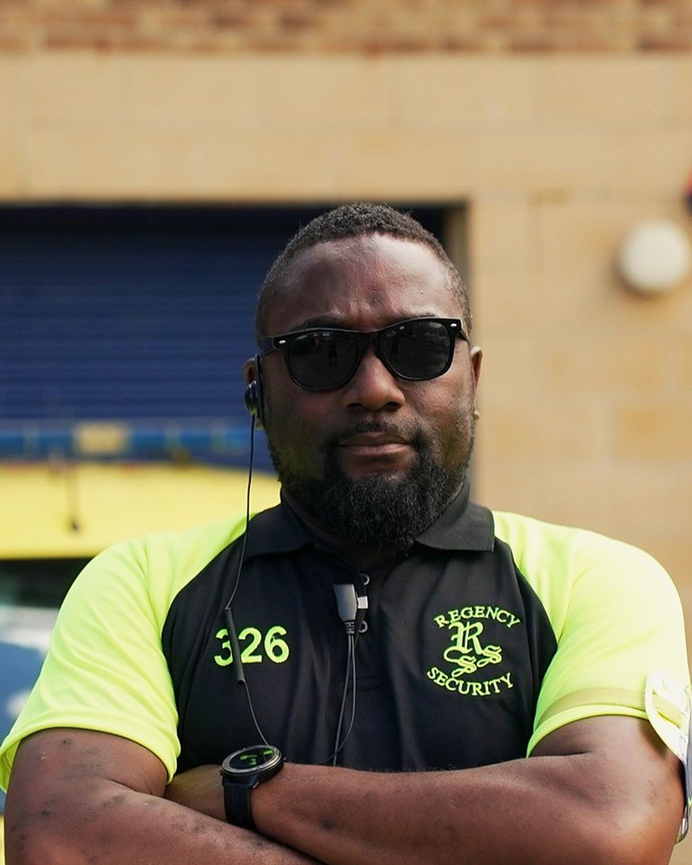 Regency Security officer in sunglasses and a hi-vis polo, arms crossed, in front of a brick building