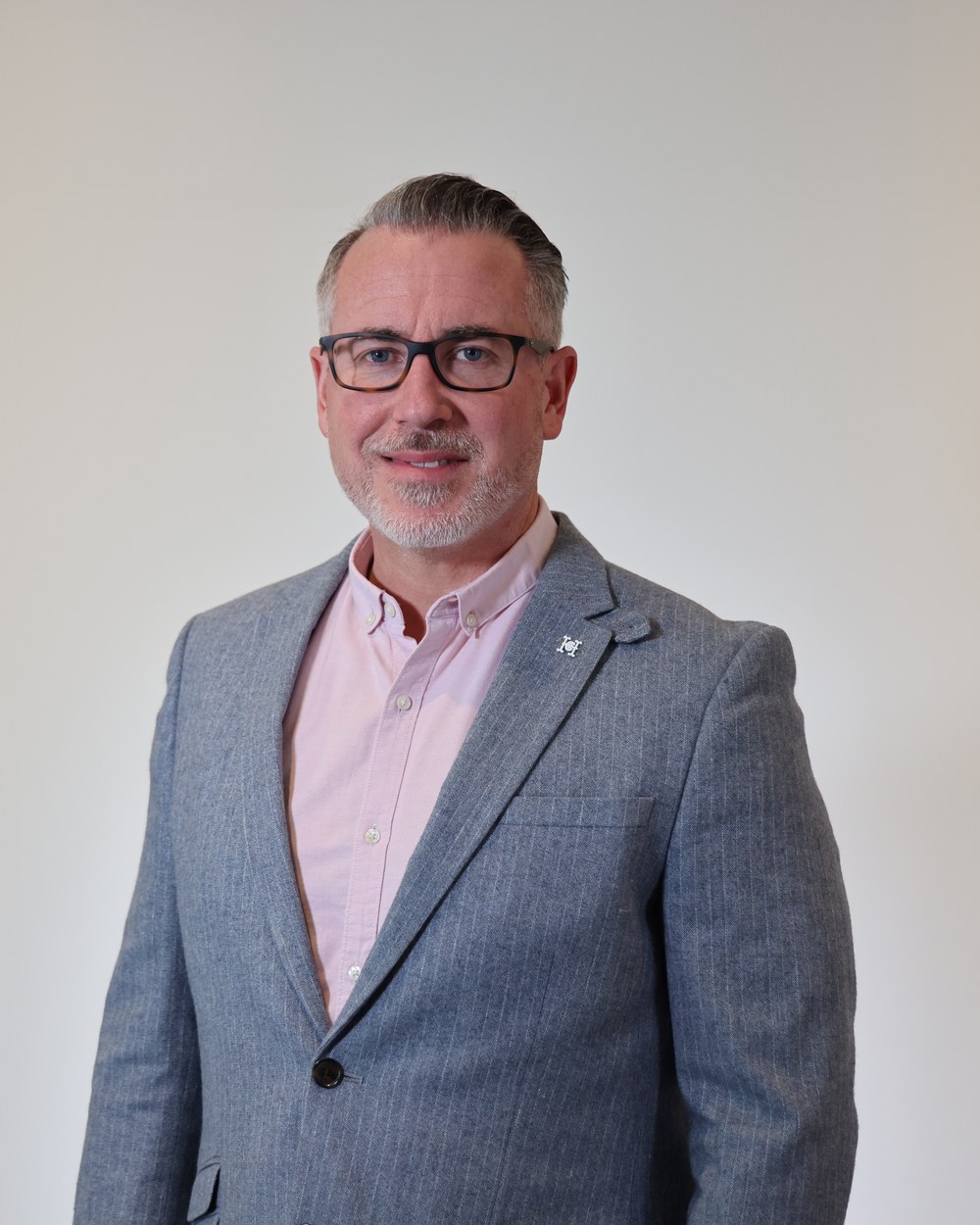 Smiling man with glasses and a grey beard in a grey blazer and pink shirt against a pale grey backdrop
